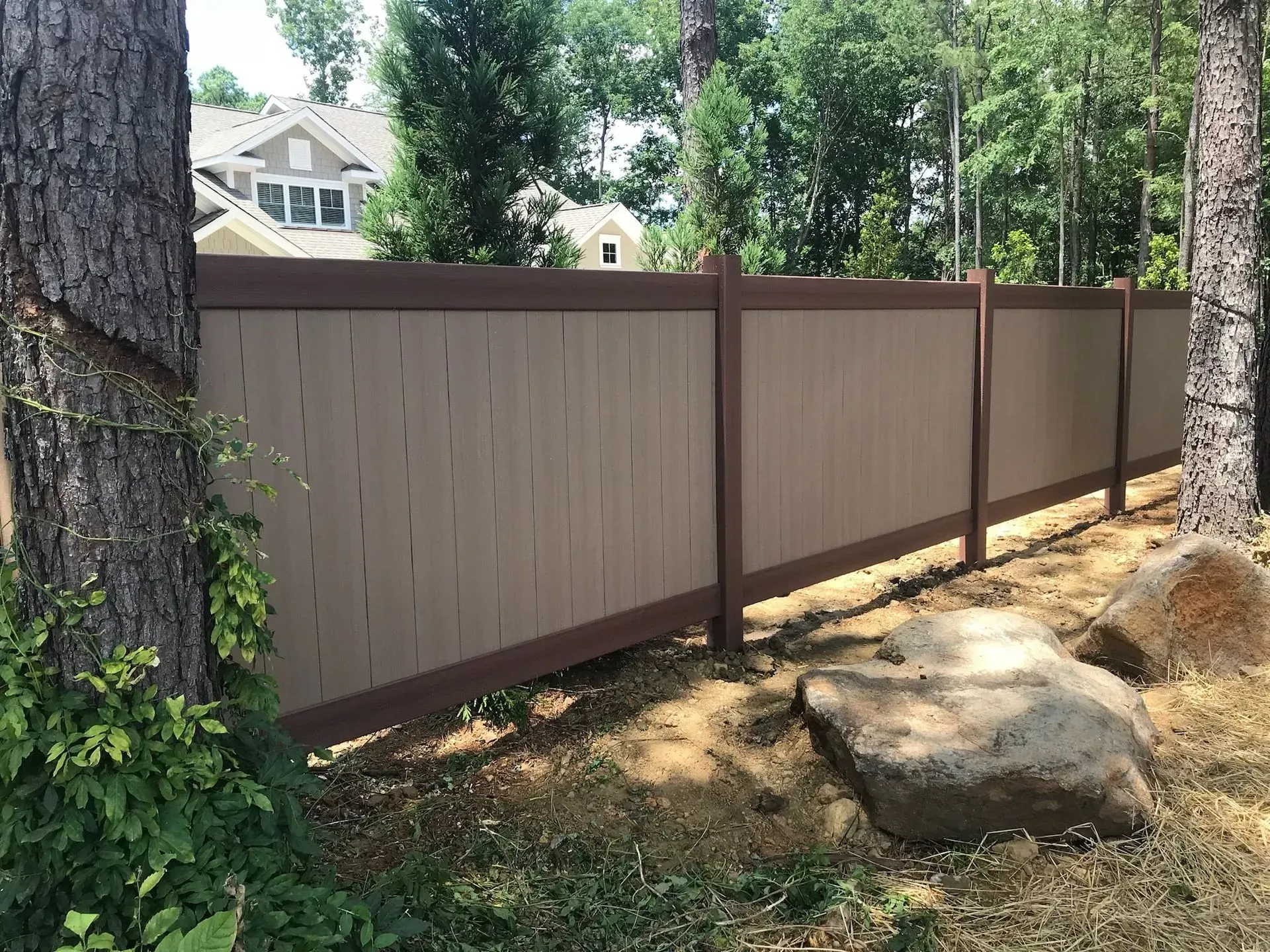 Tan composite privacy fence with brown trim in a wooded area.
