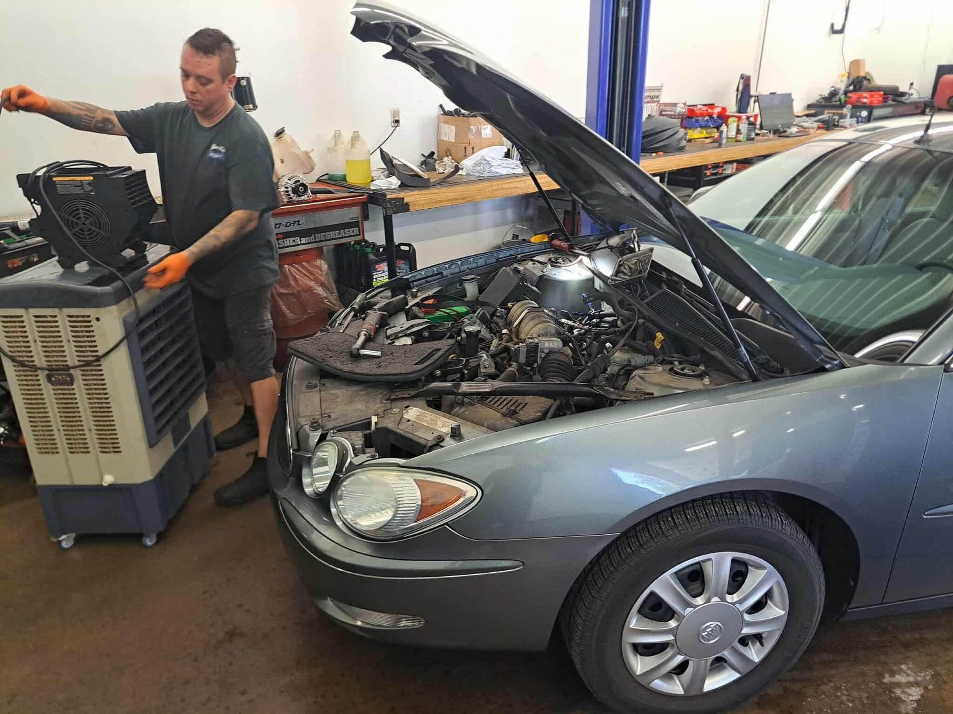 Mechanic working on a car engine with the hood open, next to an air cooler in a garage.
