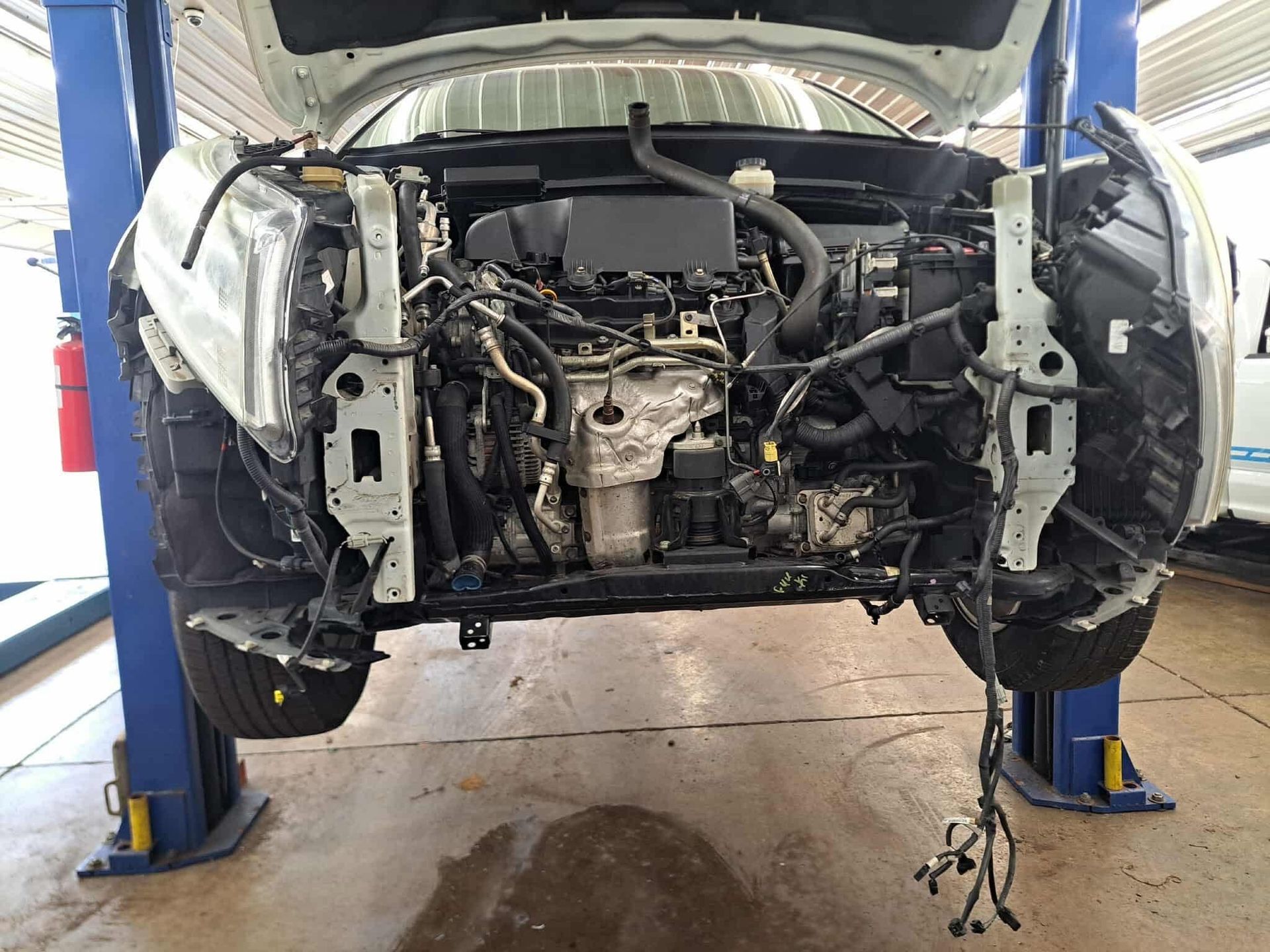 Car on a lift with the front end disassembled, revealing the engine and wiring in a repair shop.