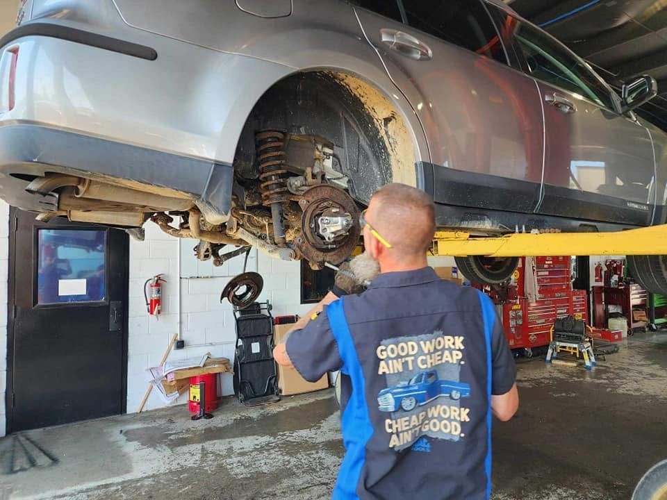 Mechanic working on a car raised on a lift, in a garage. The mechanic wears a shirt that says,