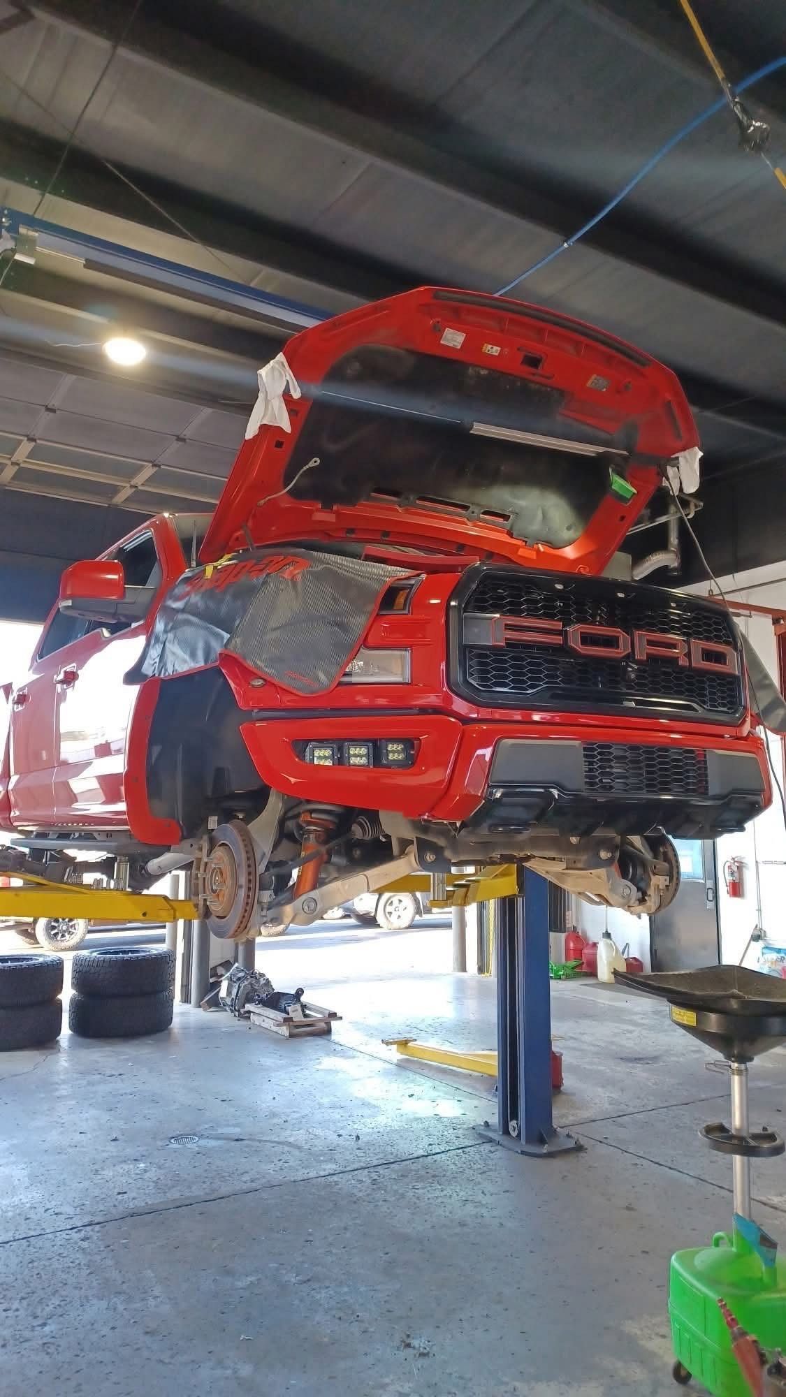 Red Ford pickup truck on a lift in a garage, hood open, undergoing repairs.