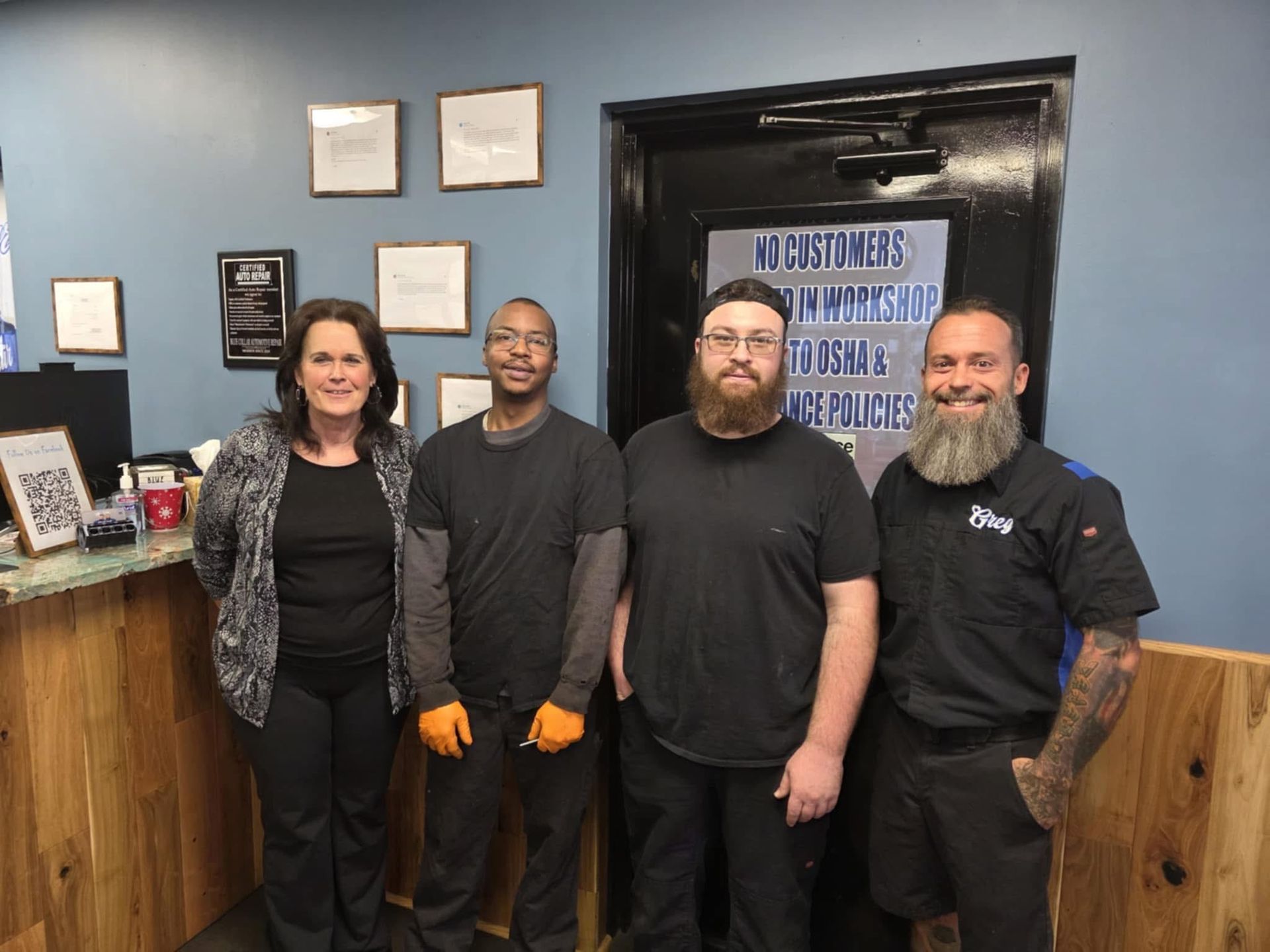 Four people standing inside an auto shop. Two men have beards, blue-collar attire. A woman smiles, dressed in black.