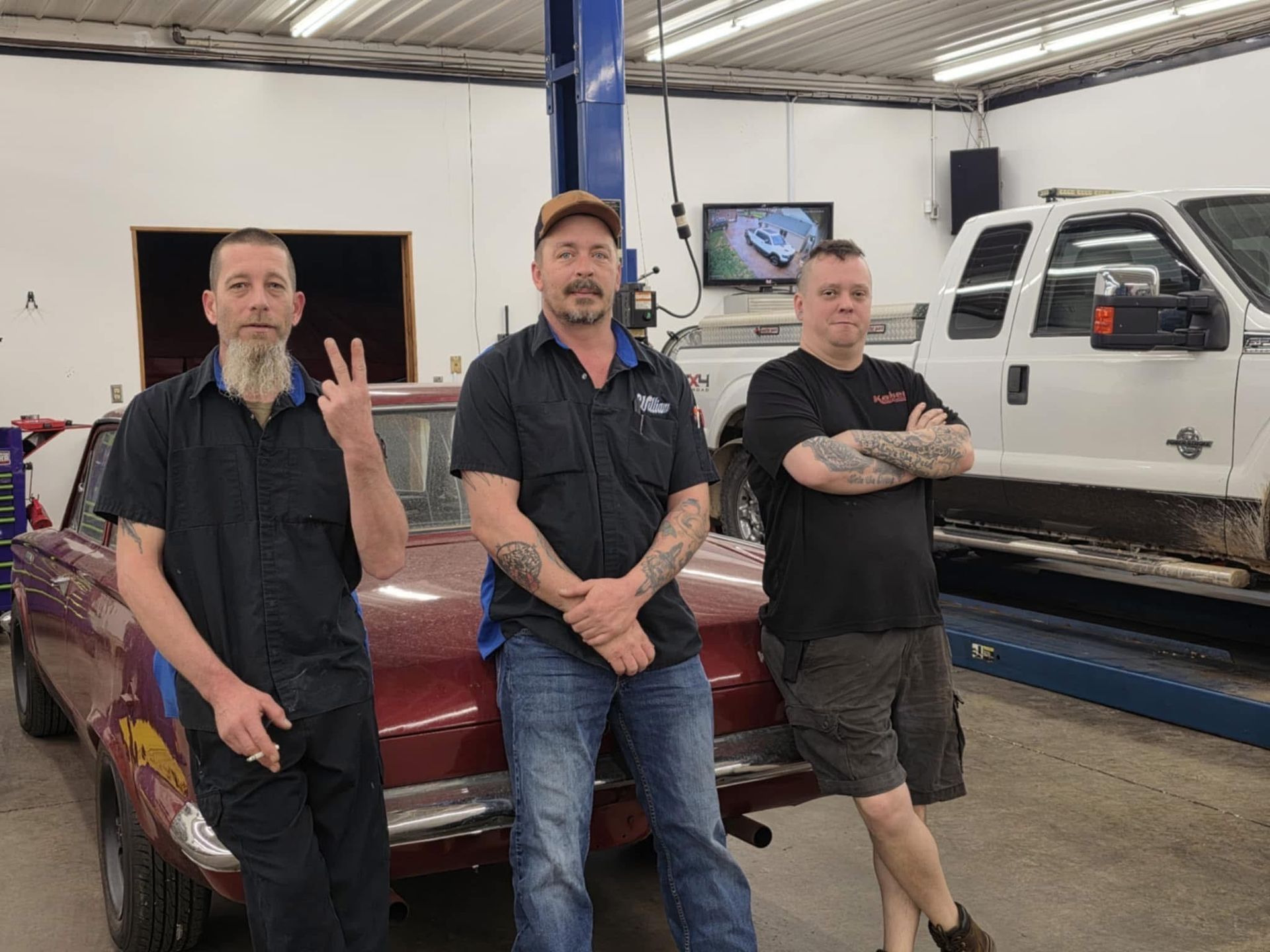 Three men pose in a garage with a classic car and truck in the background. One man makes a peace sign.