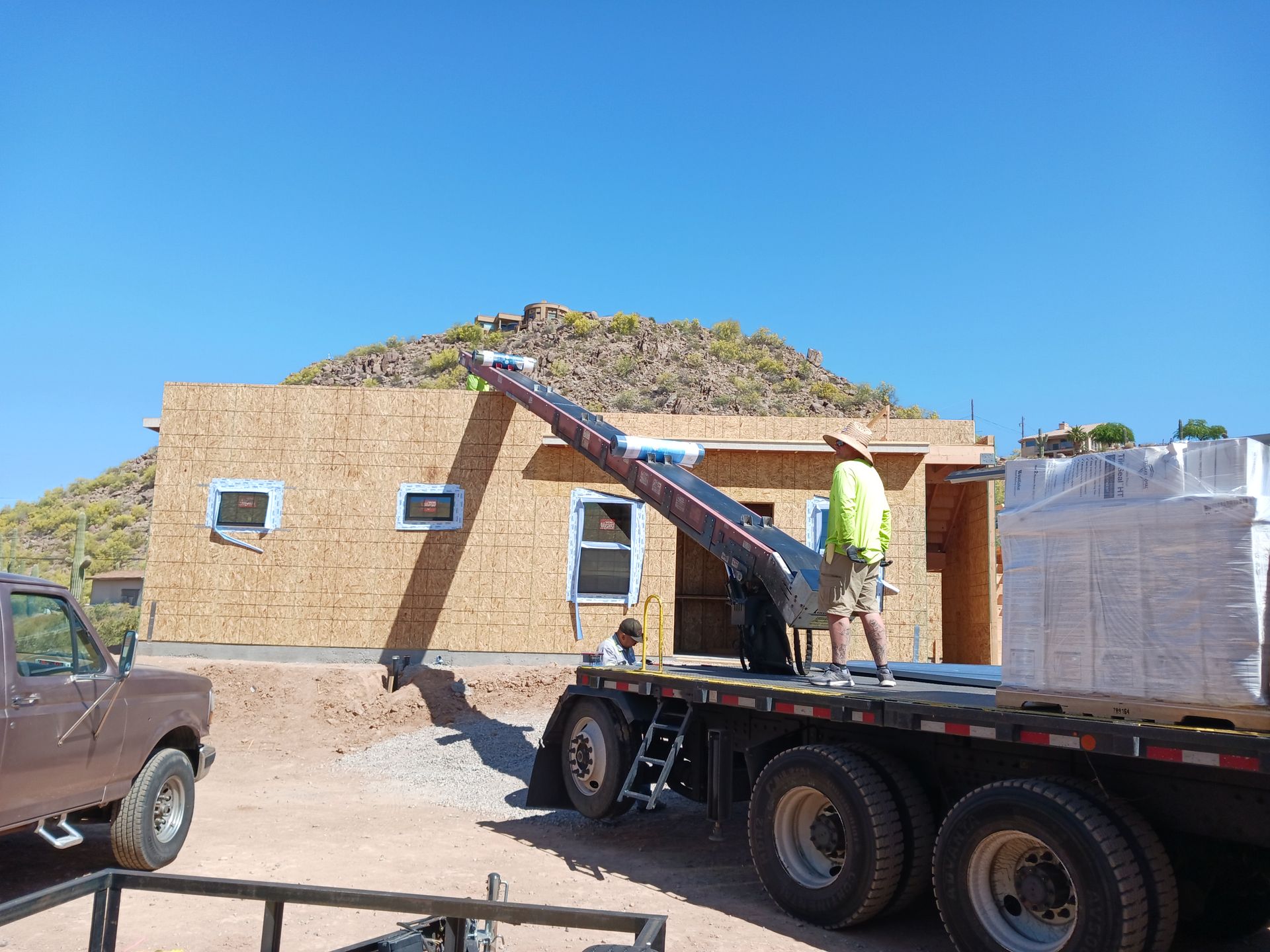 Flat-roof house under construction with a crane truck lifting materials beside stacked blocks under a blue sky