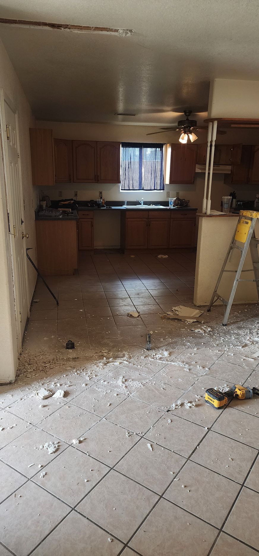 Damaged kitchen with debris on tiled floor, broken ceiling, cabinets, and window in the background