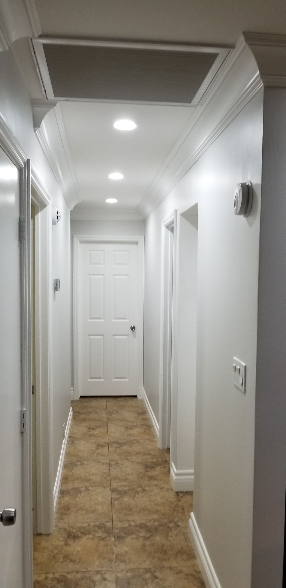 Narrow white hallway with recessed lights, beige tile floor, and a closed door at the end.