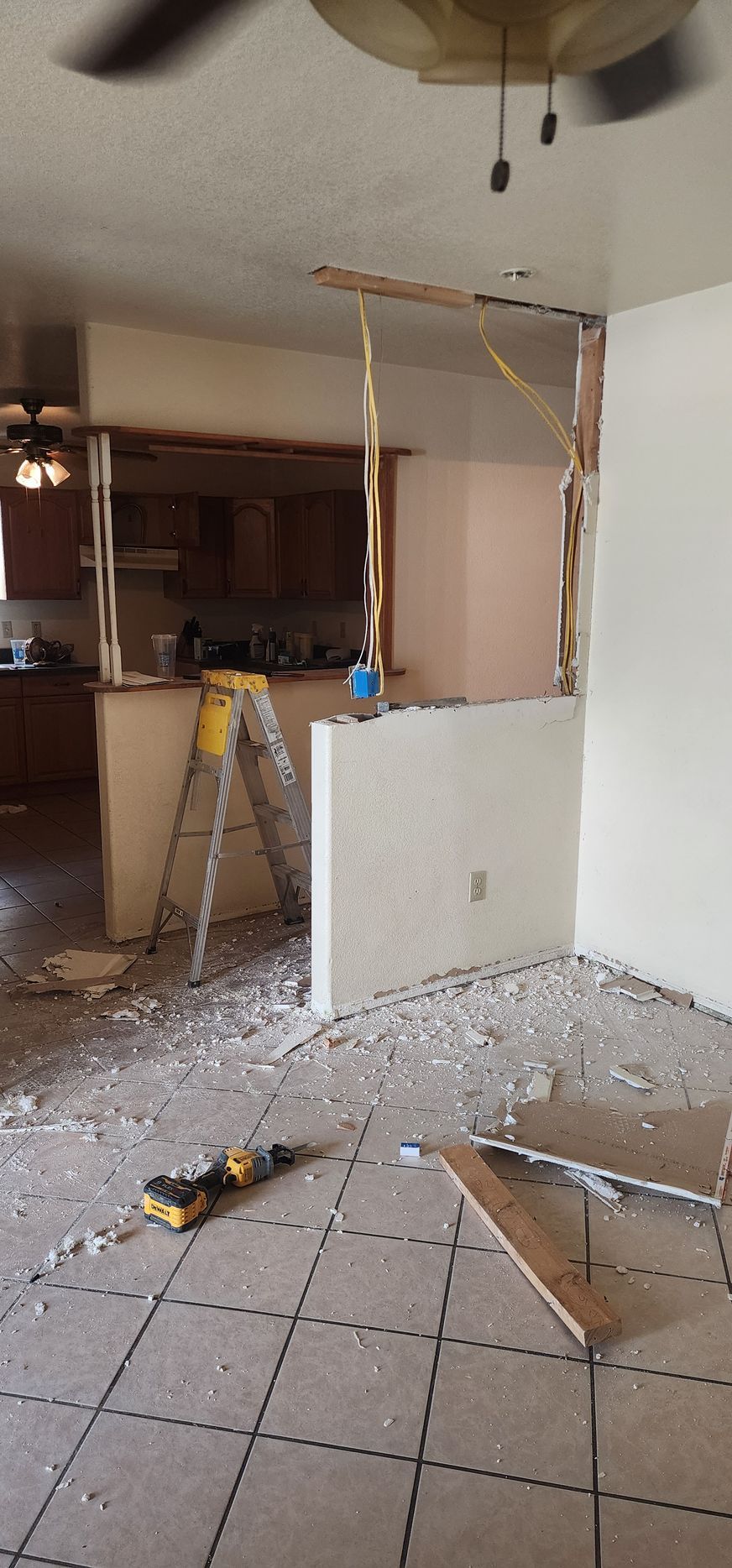 Damaged kitchen with broken wall, exposed wires, debris on tile floor, and a partially removed partition