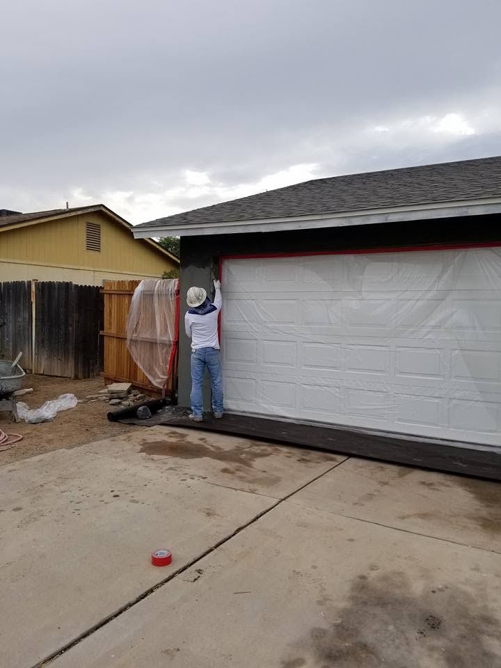 Worker paints trim on a house exterior beside a white garage door on an overcast day.