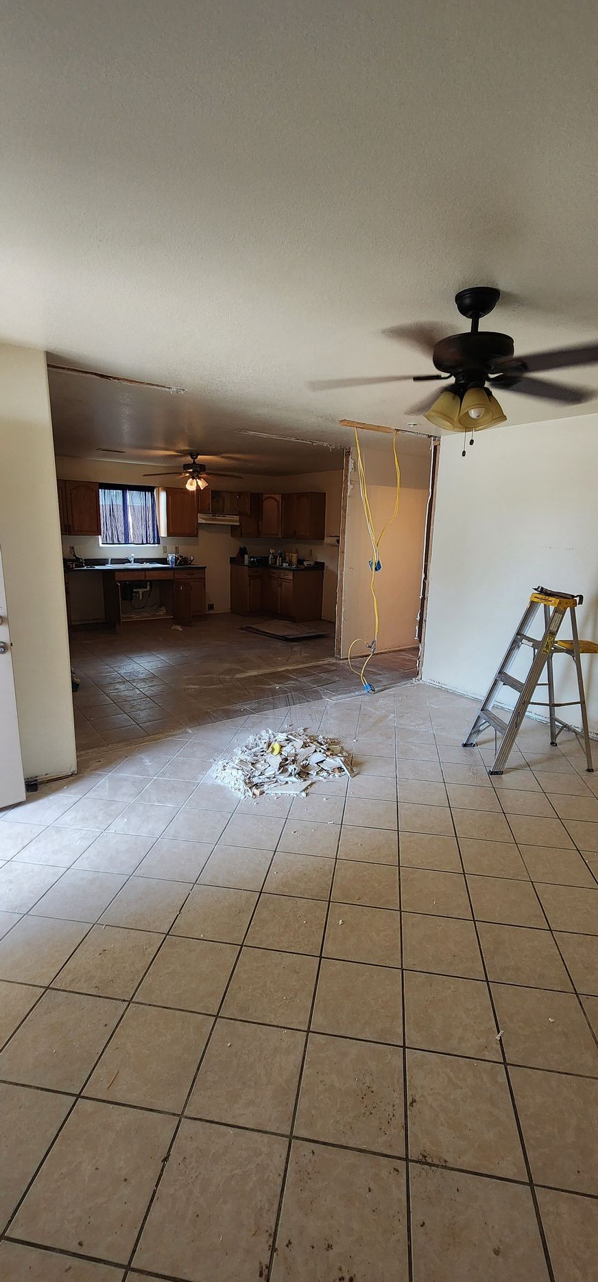 Damaged kitchen with missing ceiling and debris on tile floor, ceiling fan and ladder visible