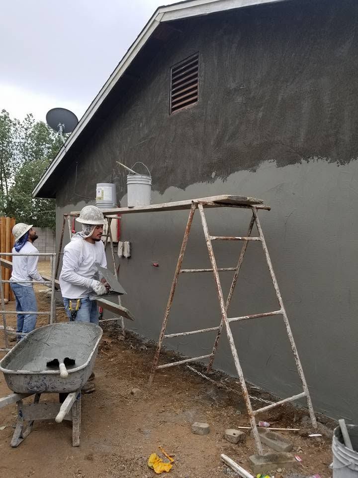 Workers plastering a gray exterior wall beside a ladder and wheelbarrow at a construction site
