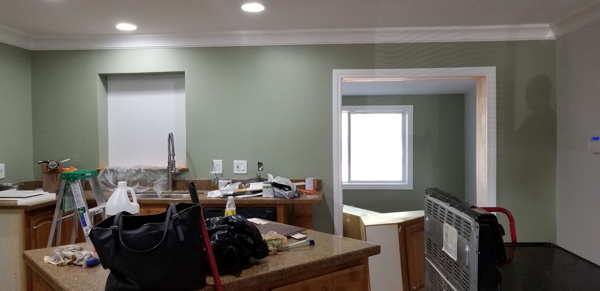 Cluttered kitchen with green walls, island, and doorway to a bright adjacent room