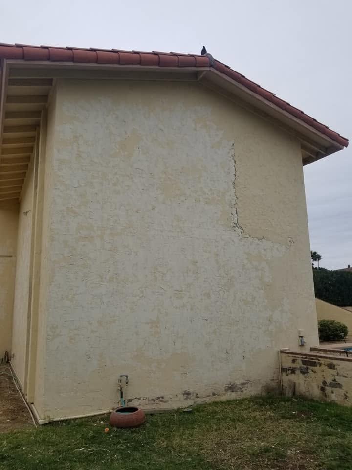 Exterior wall of a beige house with red roof tiles, showing visible cracks and peeling paint.