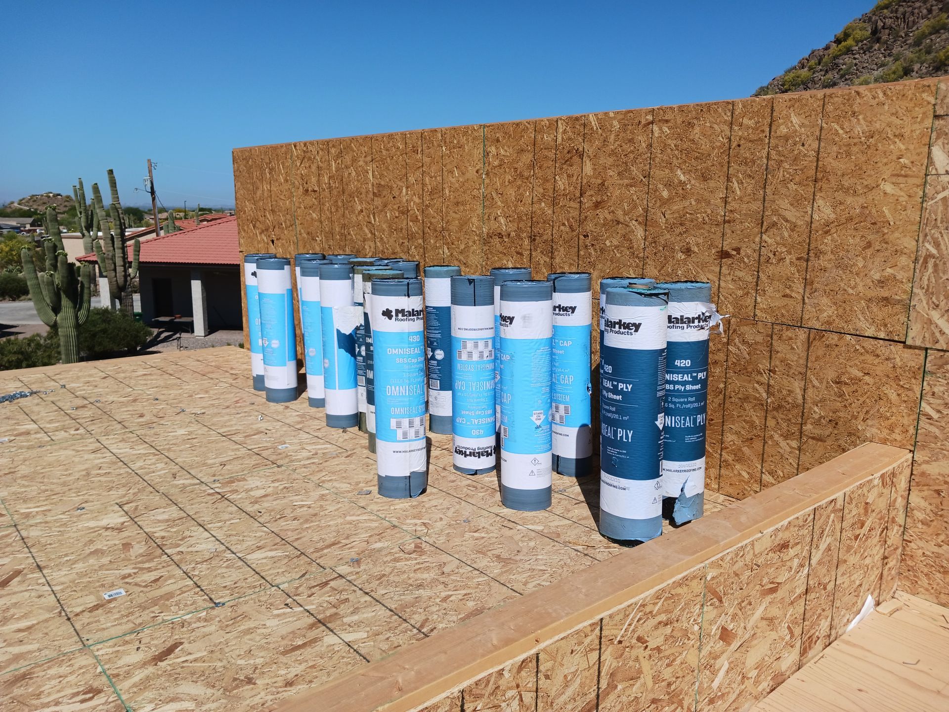 Several rolled roofing bundles on a sunlit rooftop deck beside an OSB wall under a blue sky