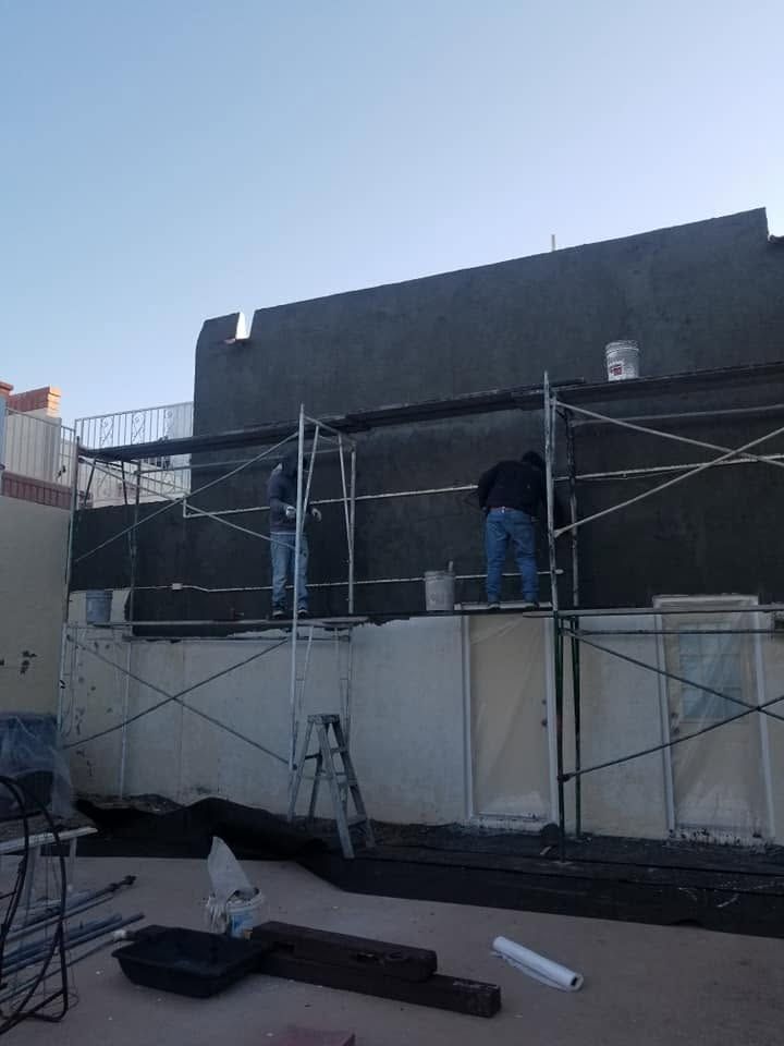 Worker on scaffolding painting a dark exterior wall beside a building under construction.
