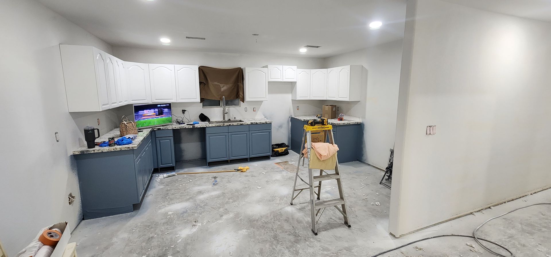 Kitchen under renovation with white cabinets, blue lower cabinets, and a step ladder on a dusty floor