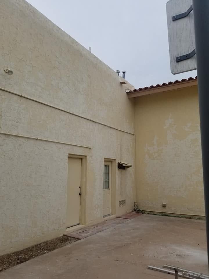 Narrow beige stucco courtyard with doors, small window, and a tiled roof under an overcast sky