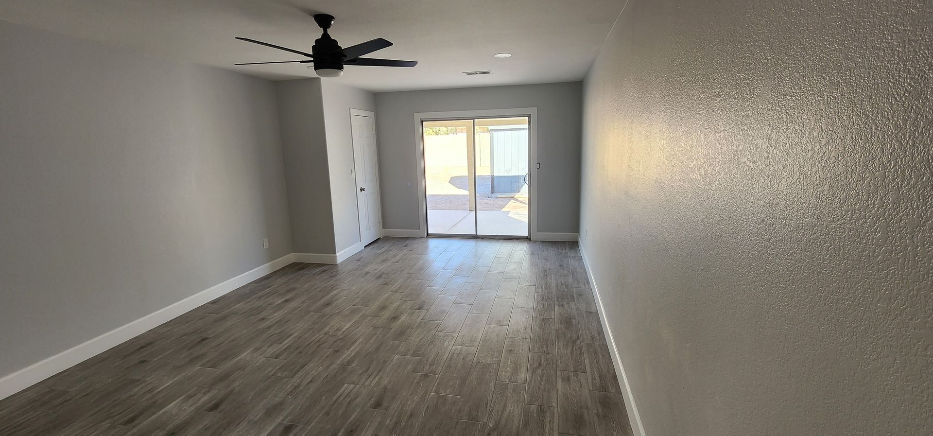 Empty living room with gray wood floors, ceiling fan, and sliding glass doors to a patio.