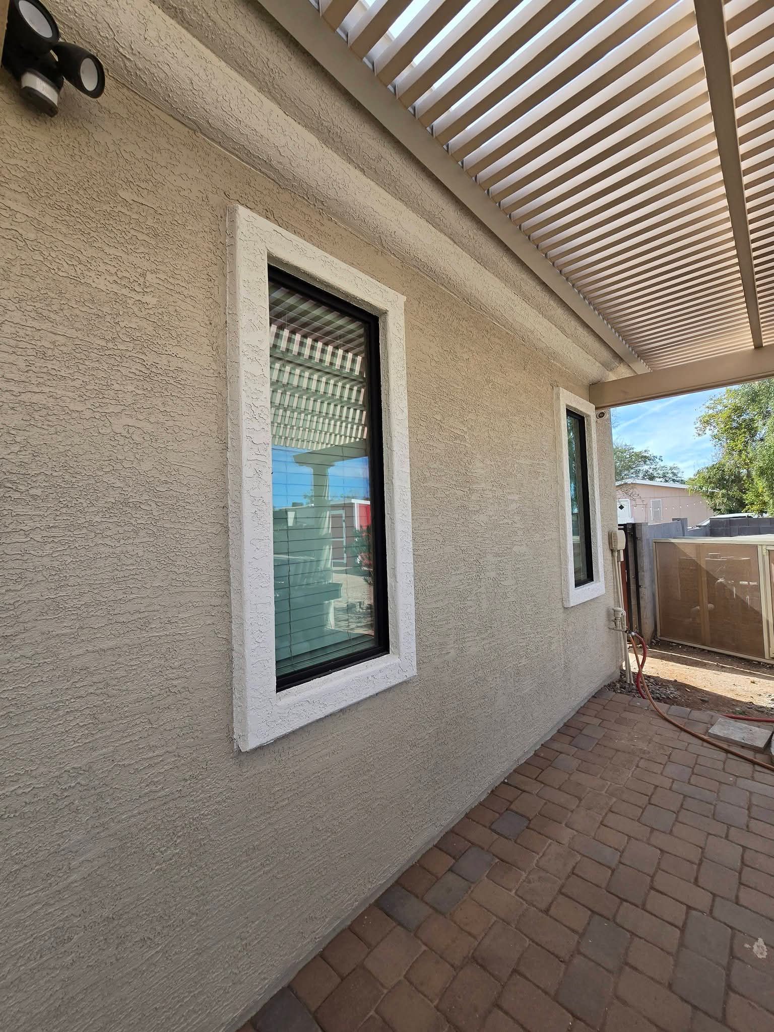 Side of a stucco house with two windows, a covered patio, and a brick walkway in sunlight.