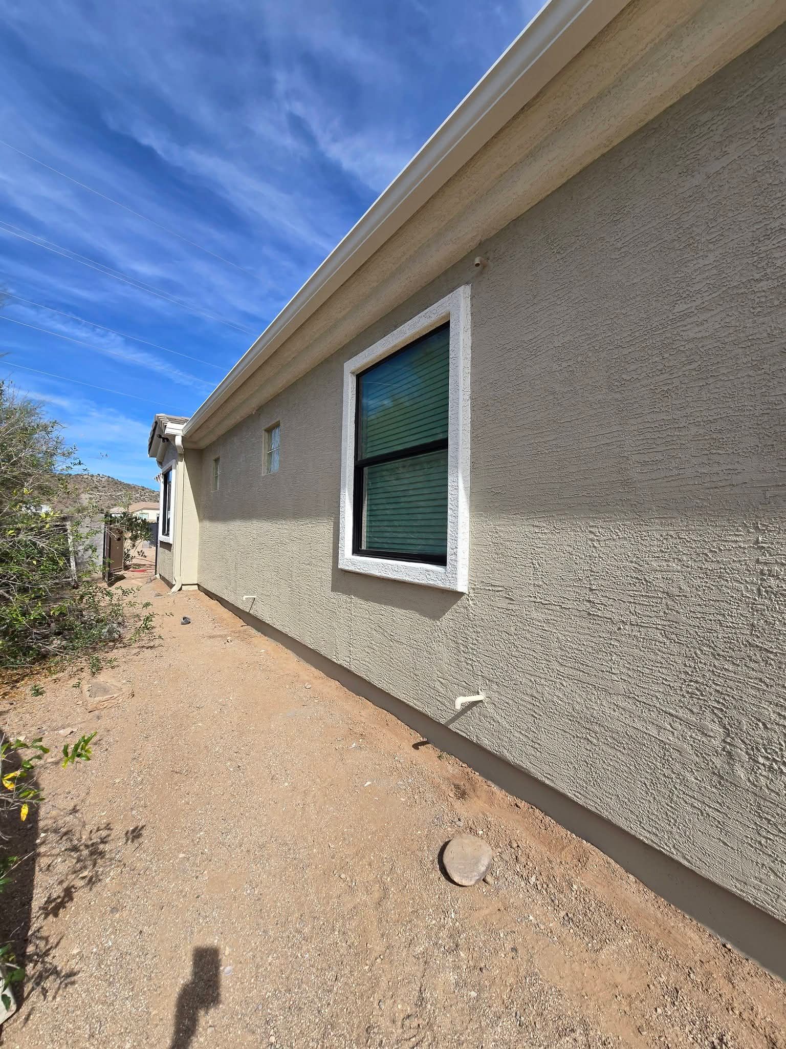 Side view of a stucco house with a green-trimmed window beside a gravel path under a blue sky.