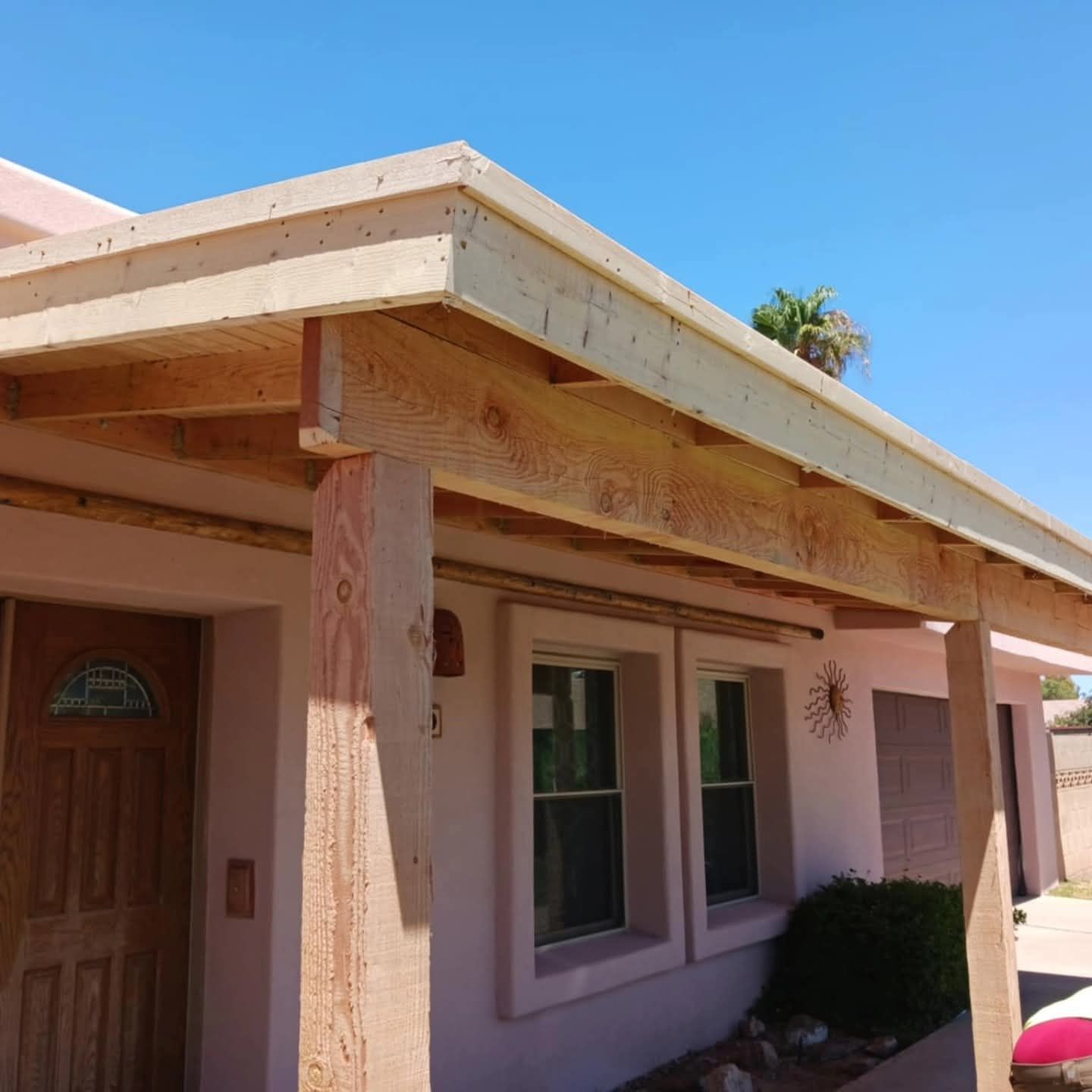 Front porch of a pink stucco house with wooden beams, a brown door, and two windows under a blue sky
