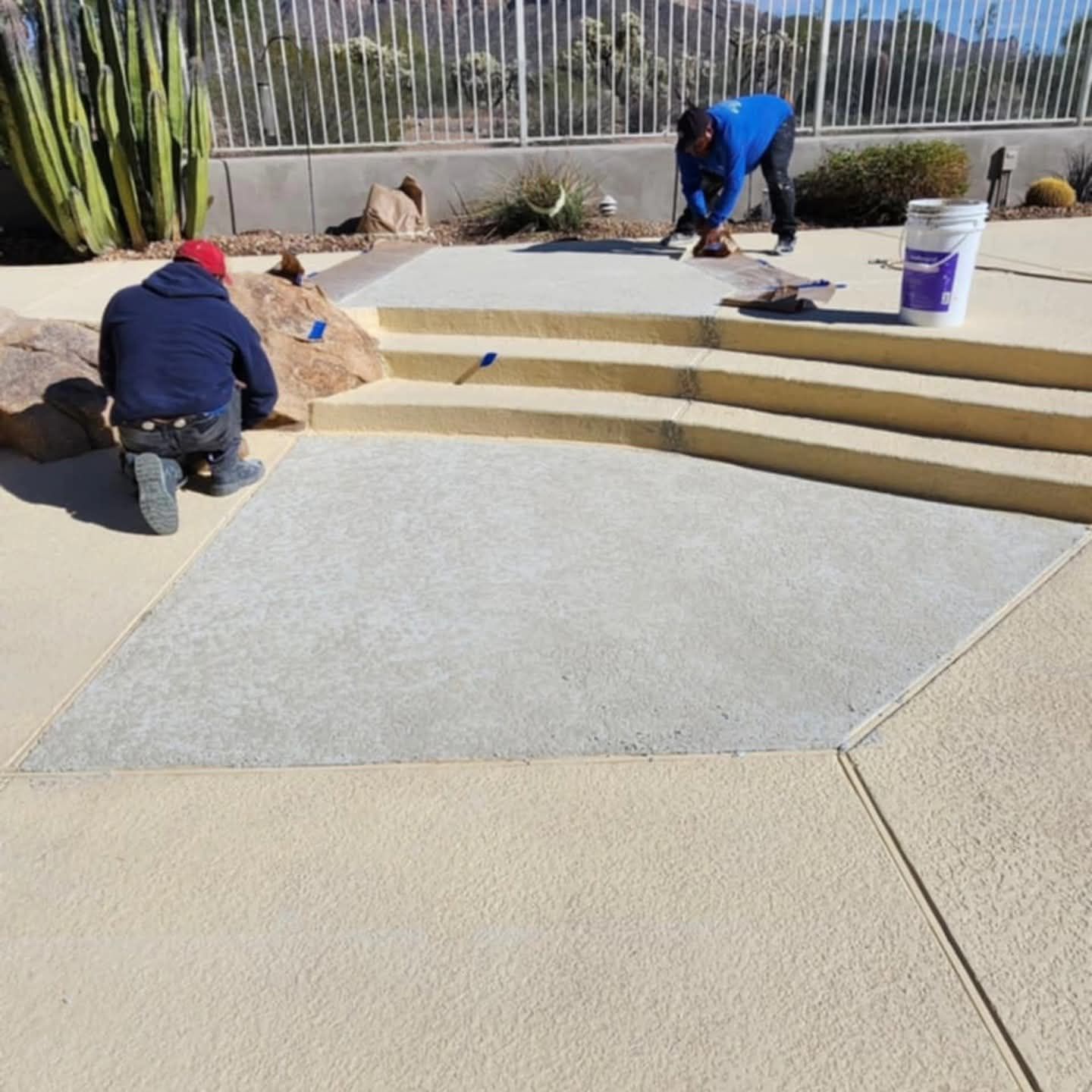 Workers applying concrete coating to a backyard patio beside curved steps and desert landscaping
