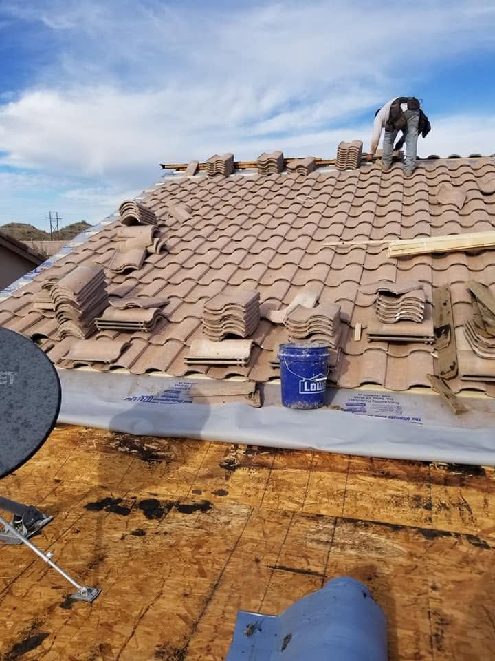 Roof under repair with worker on tan shingles, blue bucket, and exposed plywood at the edge