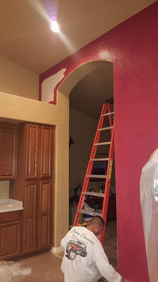 Worker on a red ladder painting a maroon arched wall indoors, beside wooden cabinets.