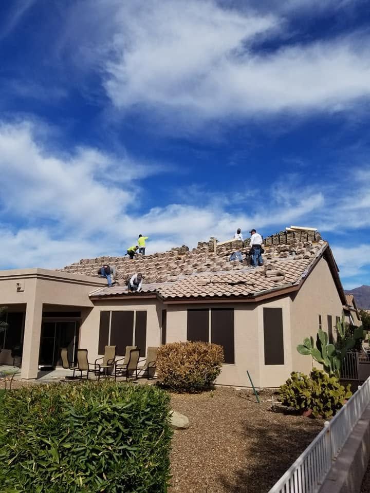 Workers repairing a tan house roof under a blue sky, with shingles and tools visible