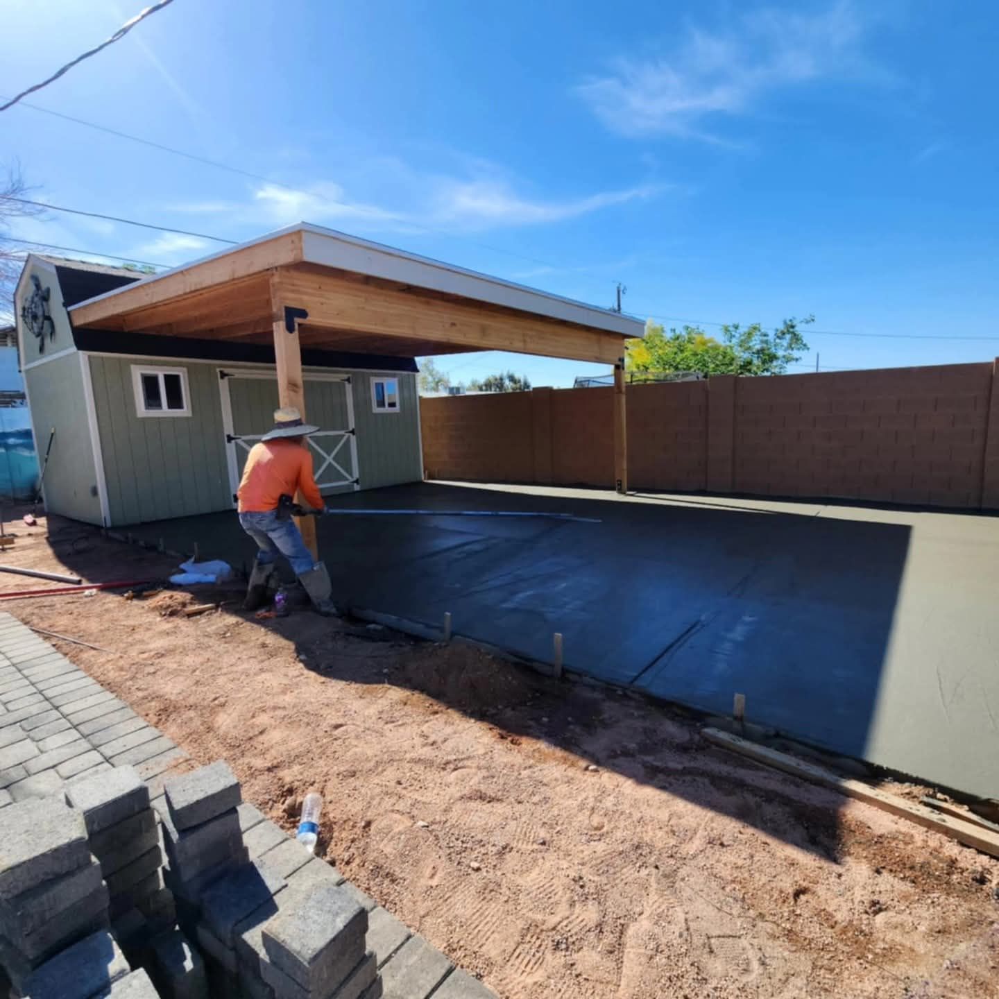 Worker smoothing fresh black driveway under a wooden carport beside a house and block wall