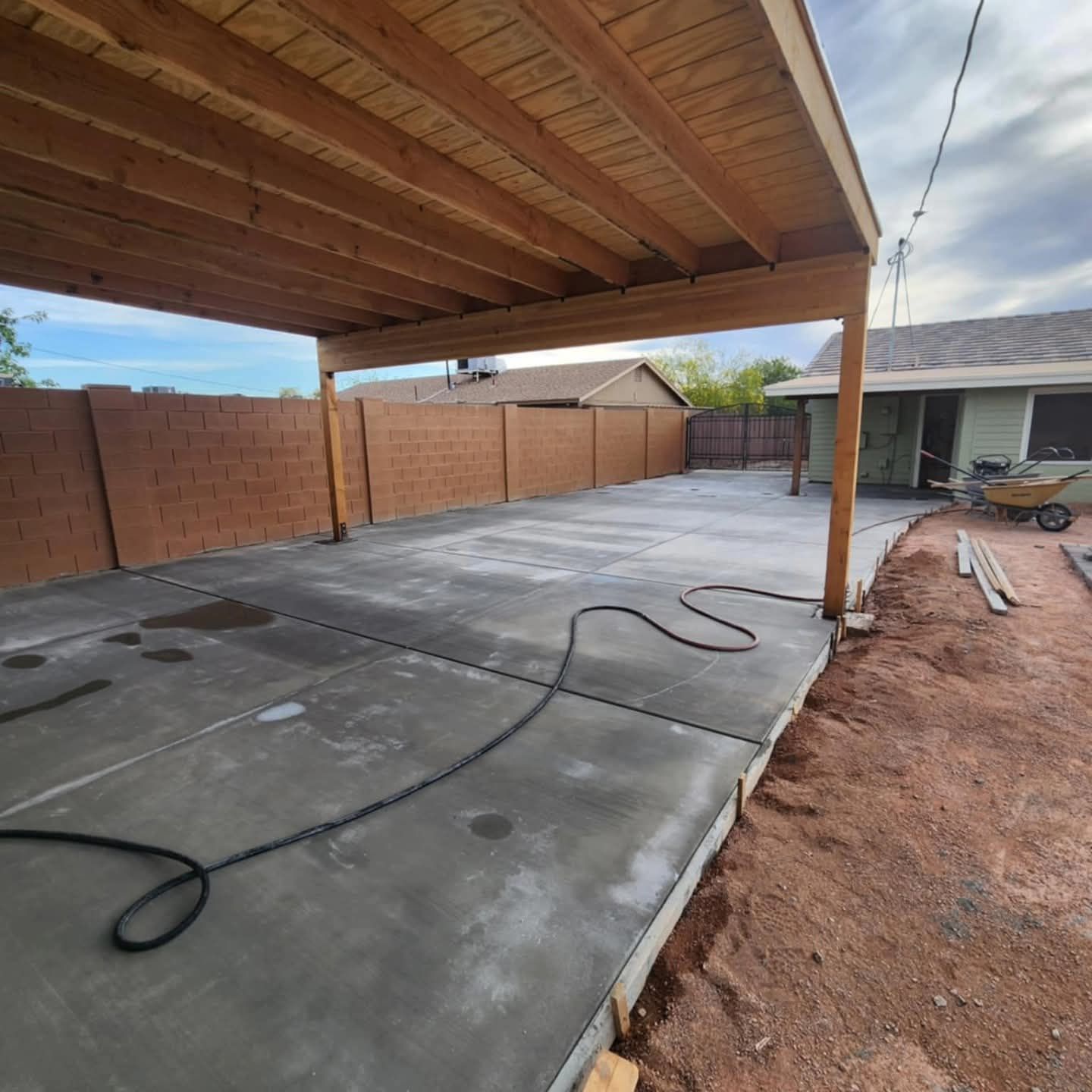 Covered concrete patio under construction with exposed wooden roof beams and a hose on the slab