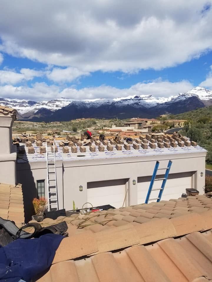 Roof work overlooking a mountain town, with ladders, terracotta tiles, and snow-capped peaks under a cloudy sky