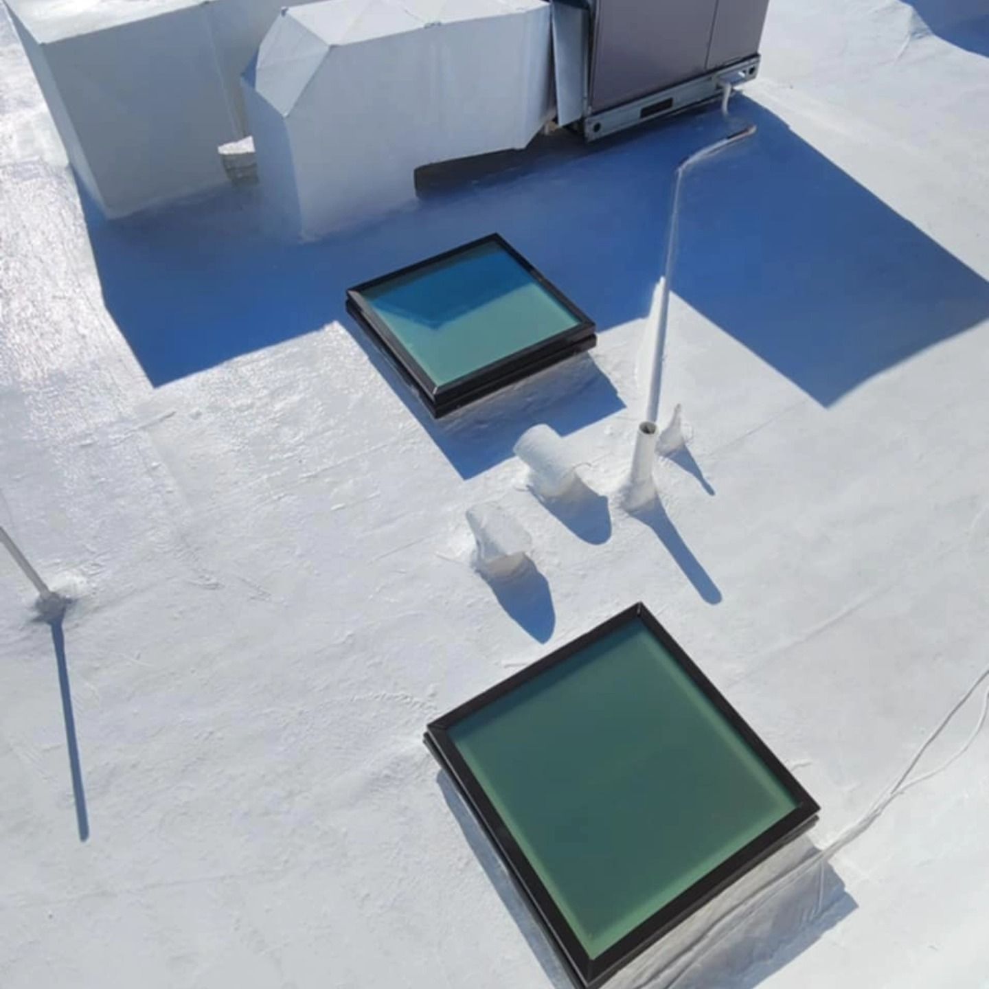 Rooftop with white surface, two square skylights, and a tall shadow from a pole near HVAC units.