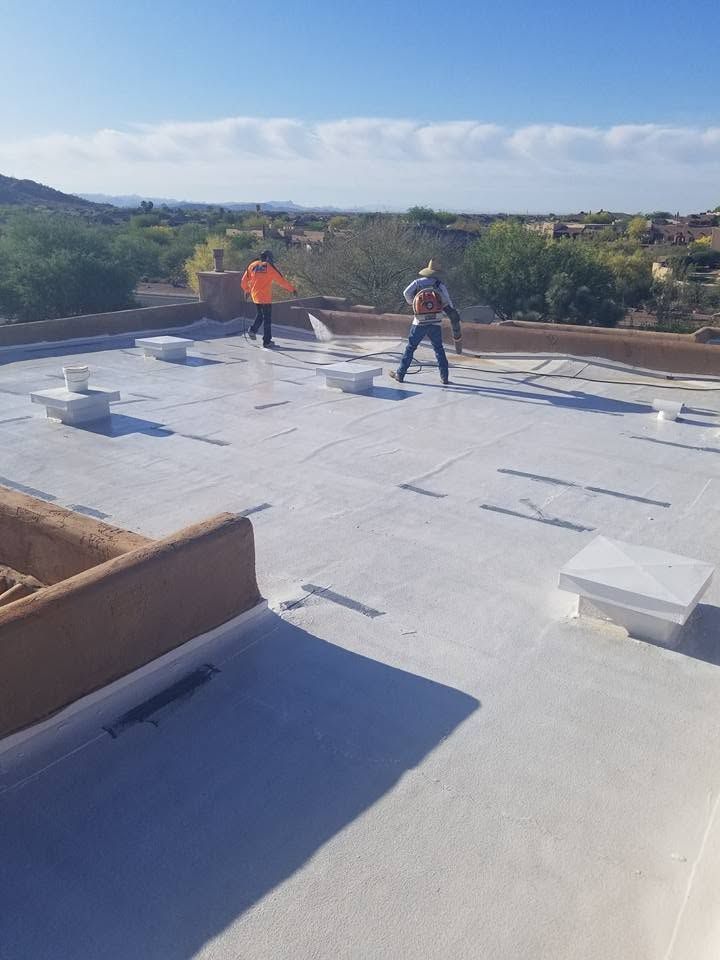 Two workers on a flat rooftop installing white roofing material under a clear sky.