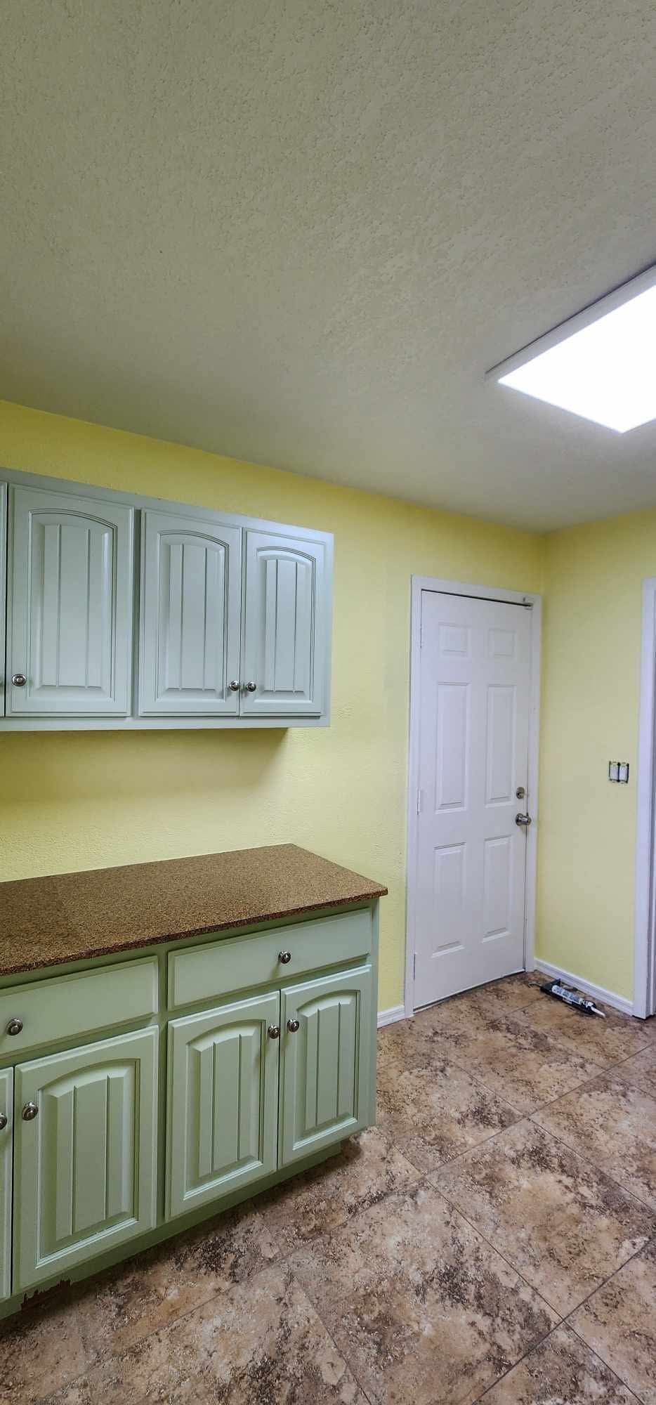 Small kitchen with mint cabinets, speckled countertop, white door, and yellow walls under bright ceiling light