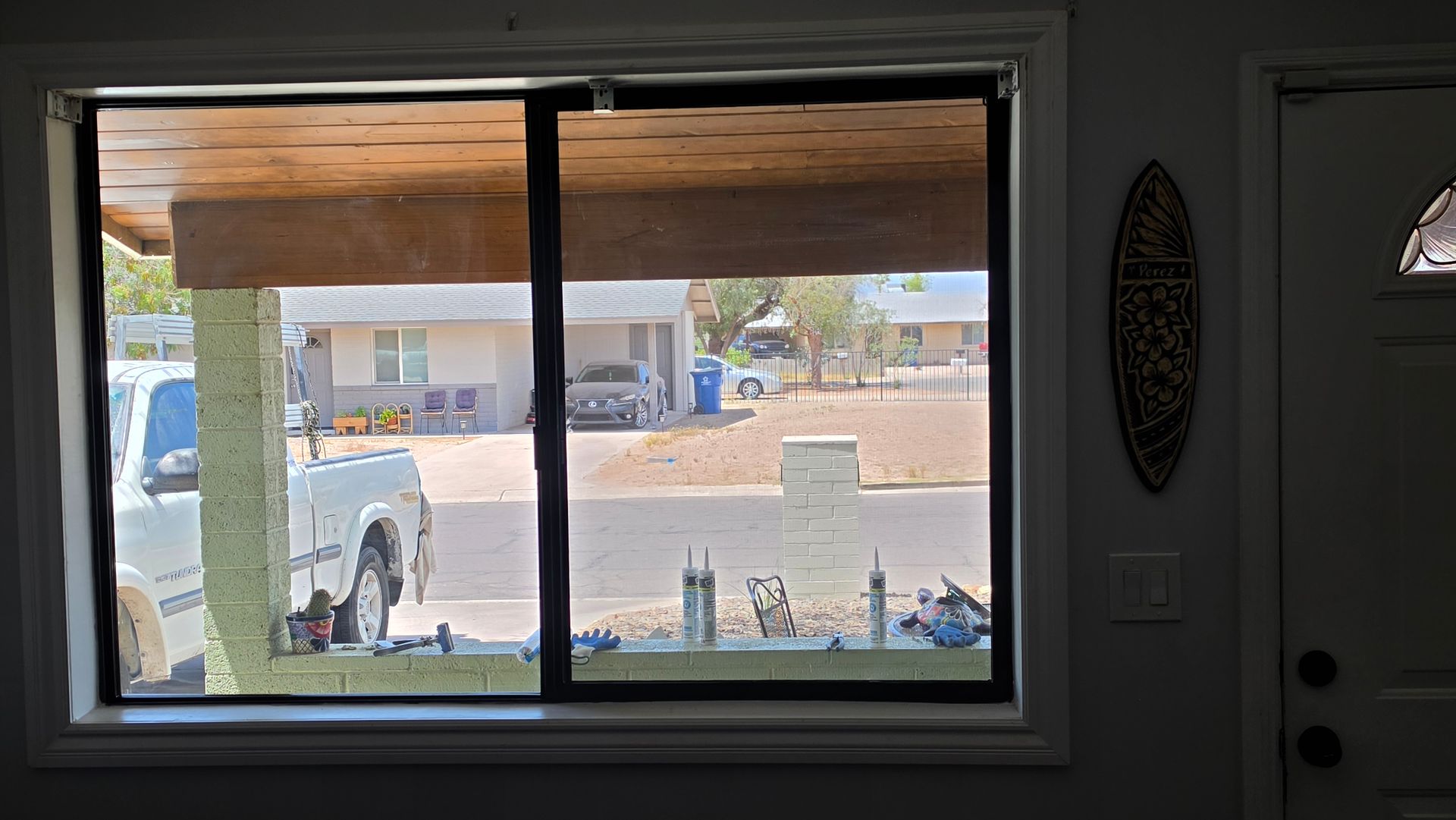 View through a living room window to a sunlit patio and street with a parked truck