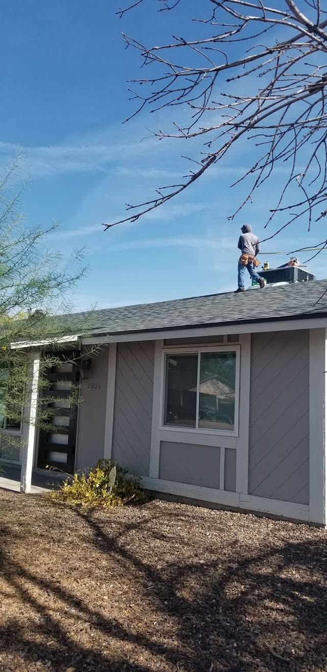 Person on a roof of a gray house under a blue sky, with trees in the foreground