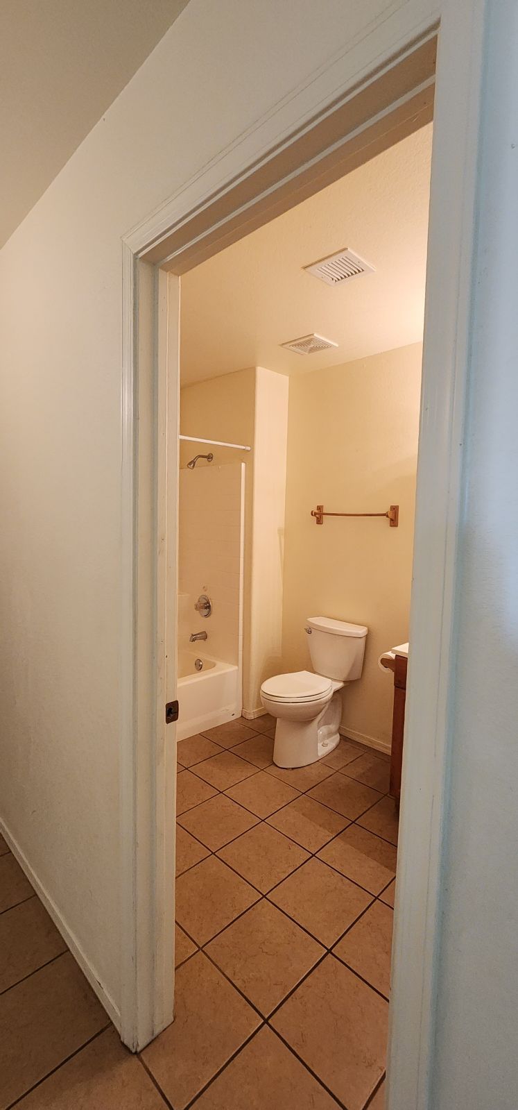 Open bathroom door with toilet, tub, towel bar, and tan tile floor inside.