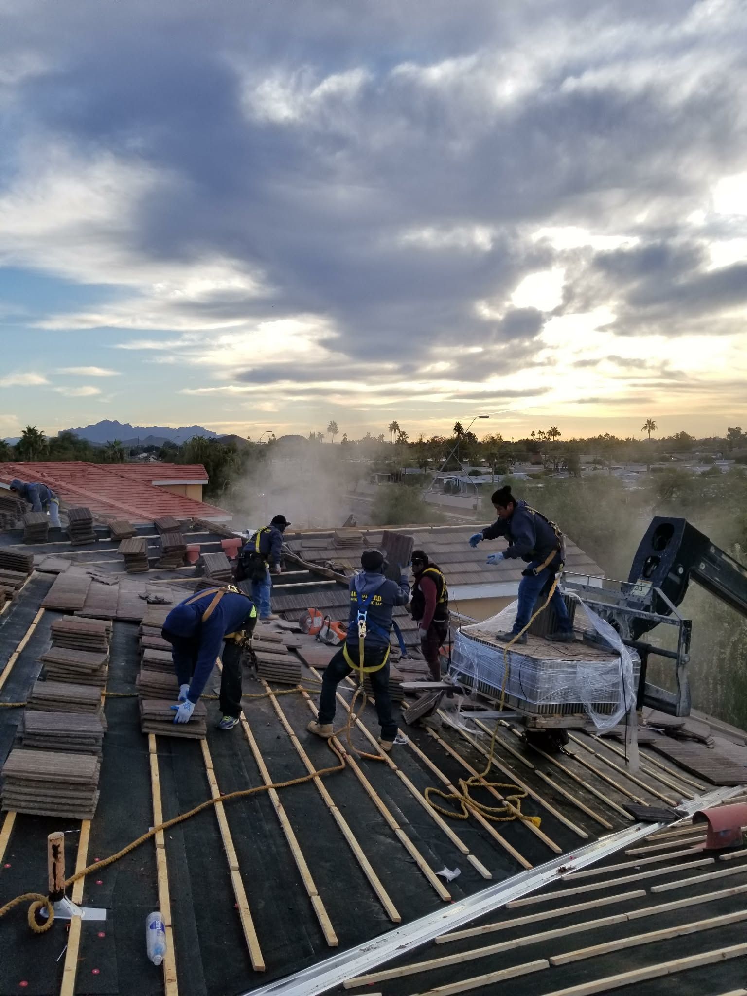 Workers pouring concrete on a rooftop at sunset, with steam or dust rising under a cloudy sky.