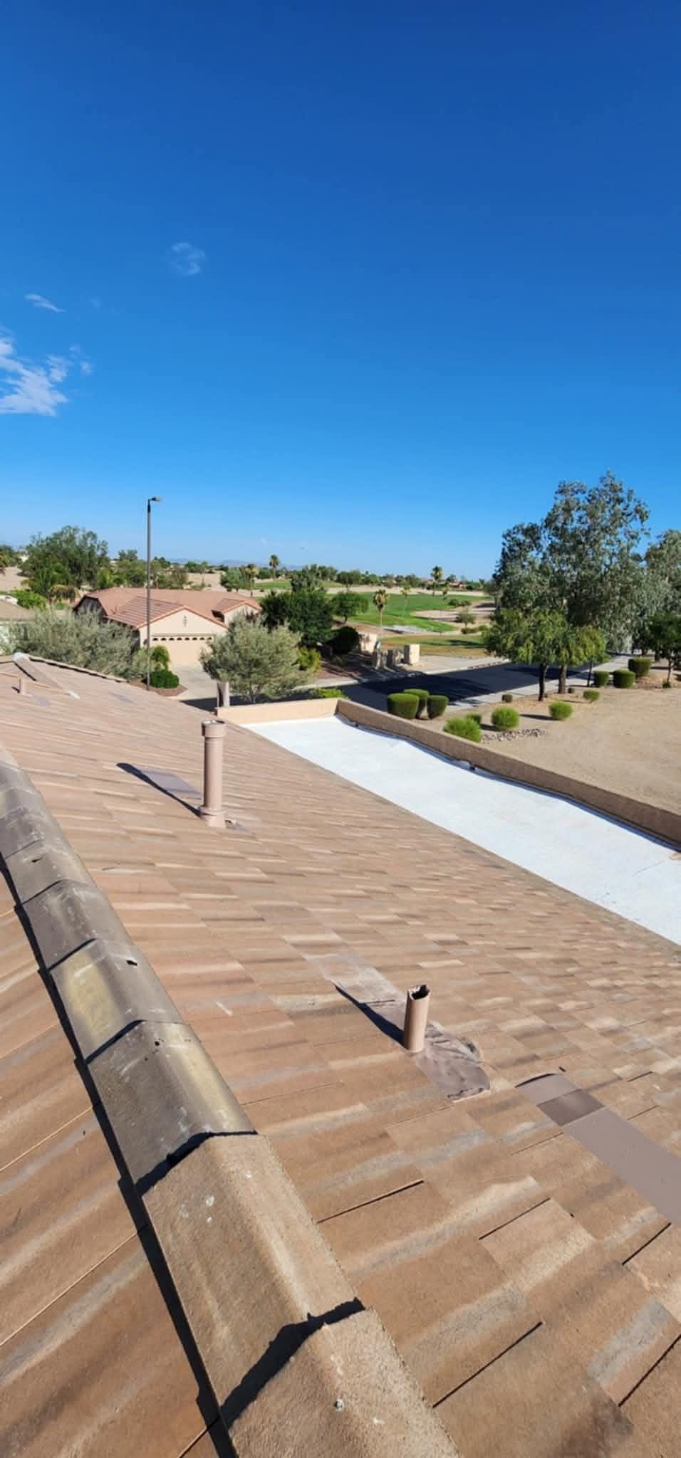 Rooftop with brown shingles, white ridge line, and a clear blue sky over a suburban landscape