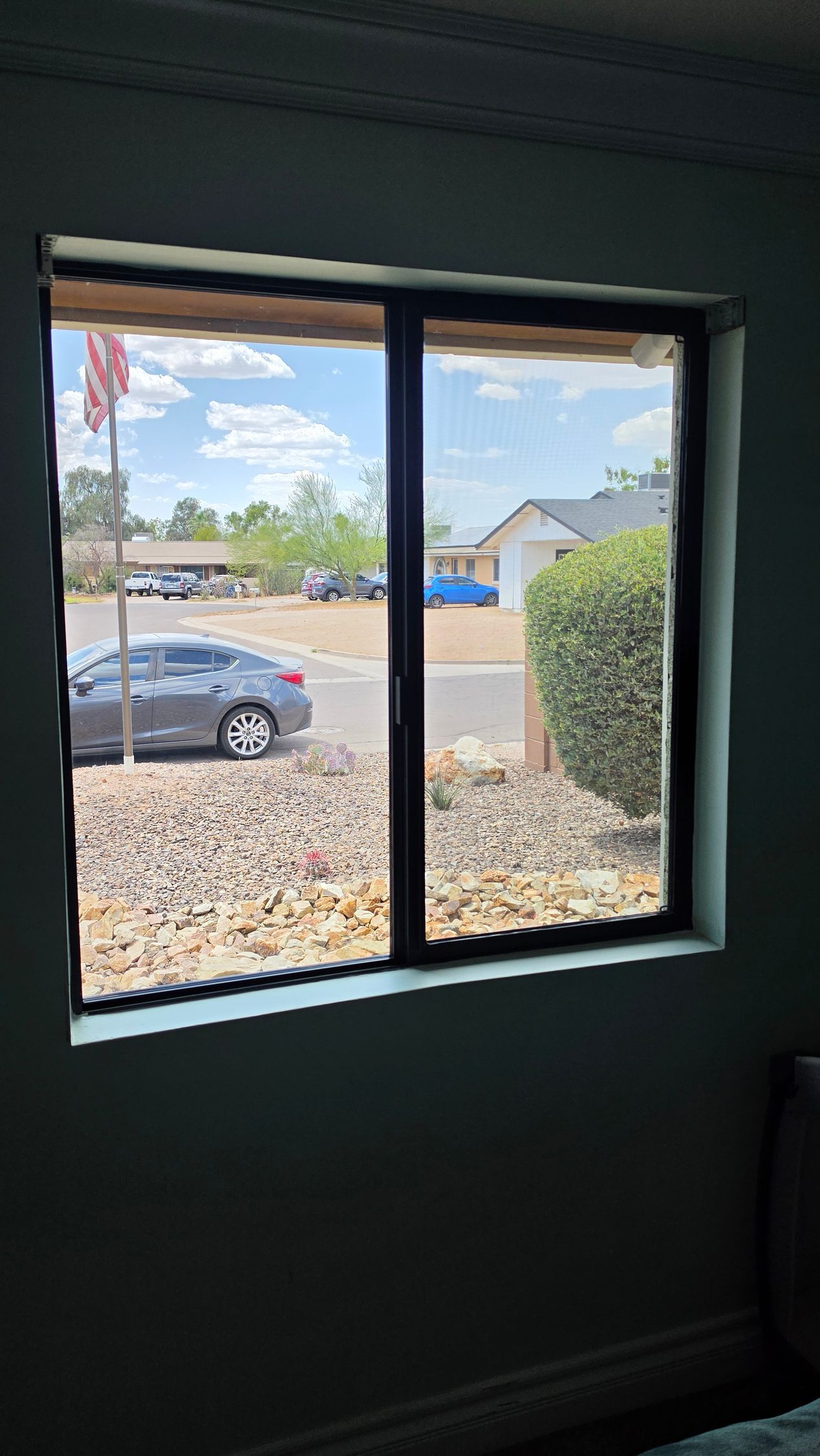 View through a dark room window of a suburban street with parked car, houses, and clear sky.