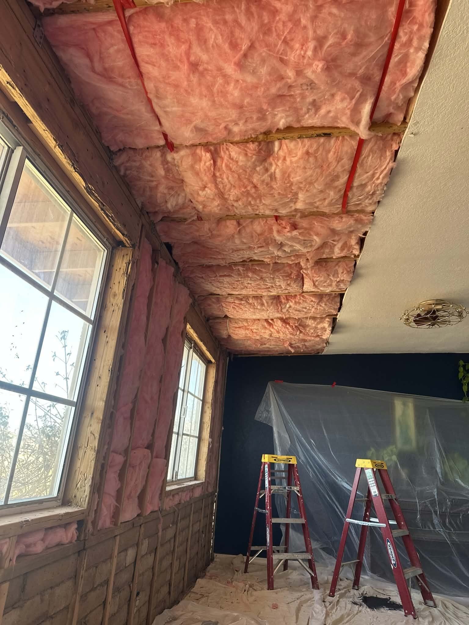 Partially renovated room with exposed pink insulation in the ceiling and ladders near a window and wall area