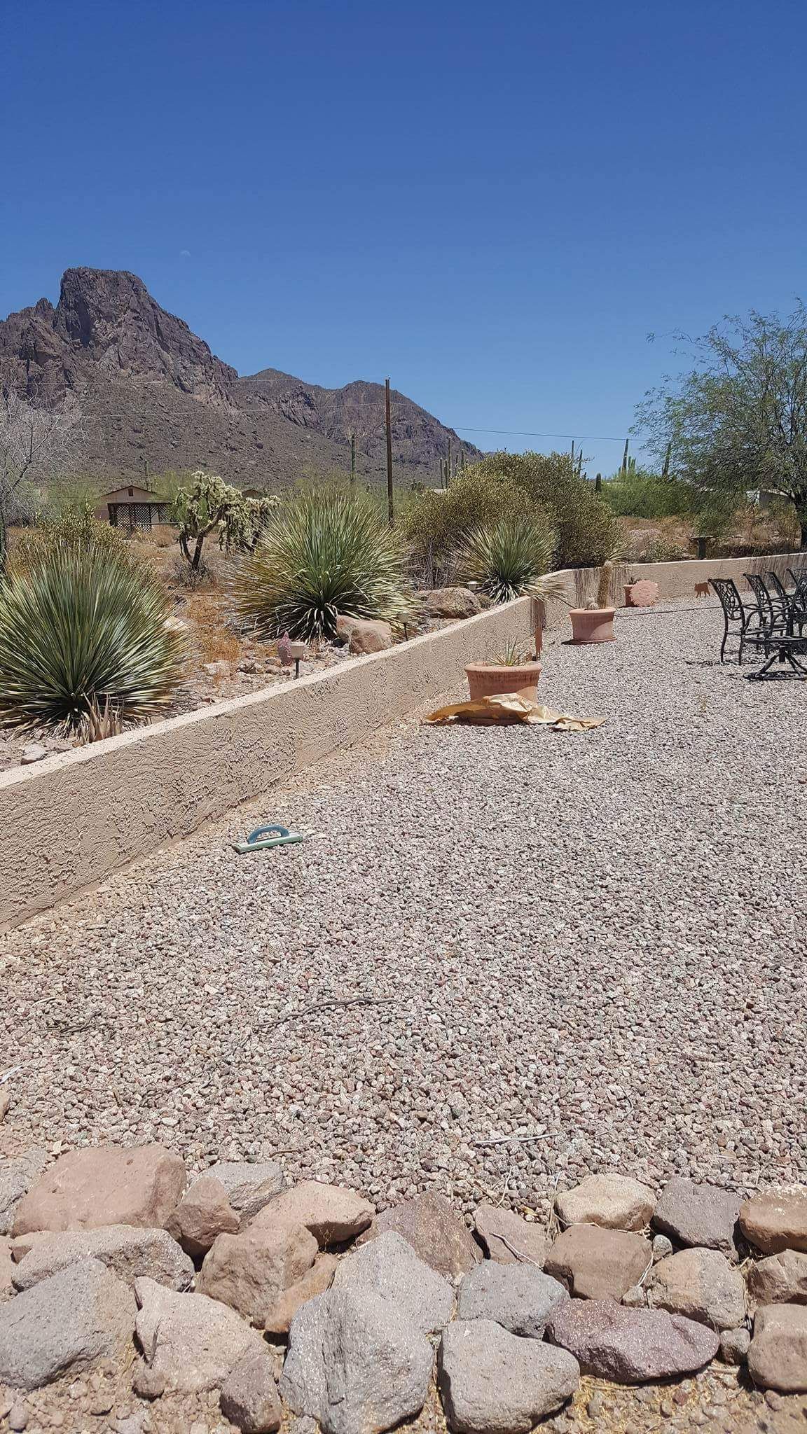 Desert landscape with gravel yard, rocks, sparse shrubs, and a mountain under a clear blue sky