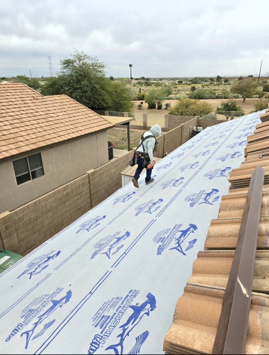 Worker installing white roof underlayment on a residential roof, viewed between tan tiled rooftops.