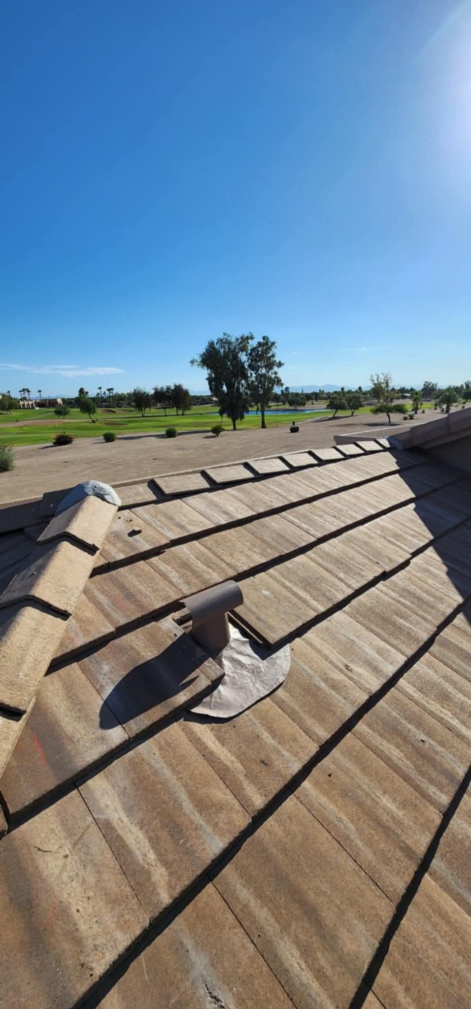Sunlit shingled roof with a vent pipe, overlooking a green field and blue sky