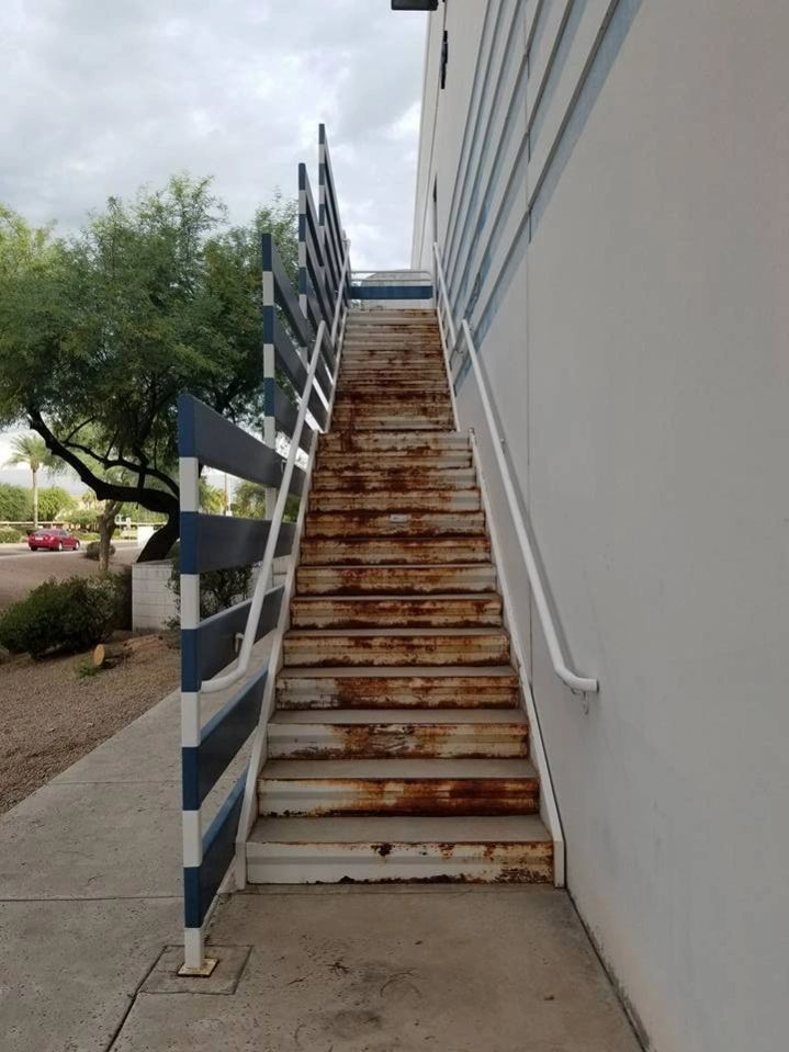 Steep outdoor metal stairway beside a white building, with striped railings and trees nearby