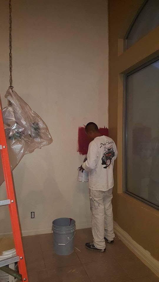Worker painting a wall beside a large window, with a ladder and bucket in a room under renovation
