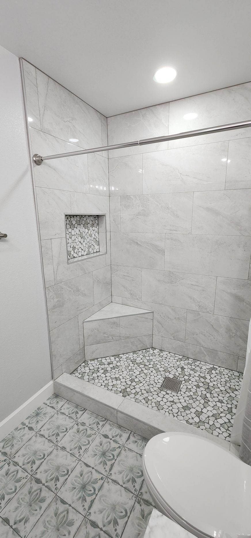 Tiled bathroom shower with glass door, pebble floor, small built-in shelf, and toilet seat visible