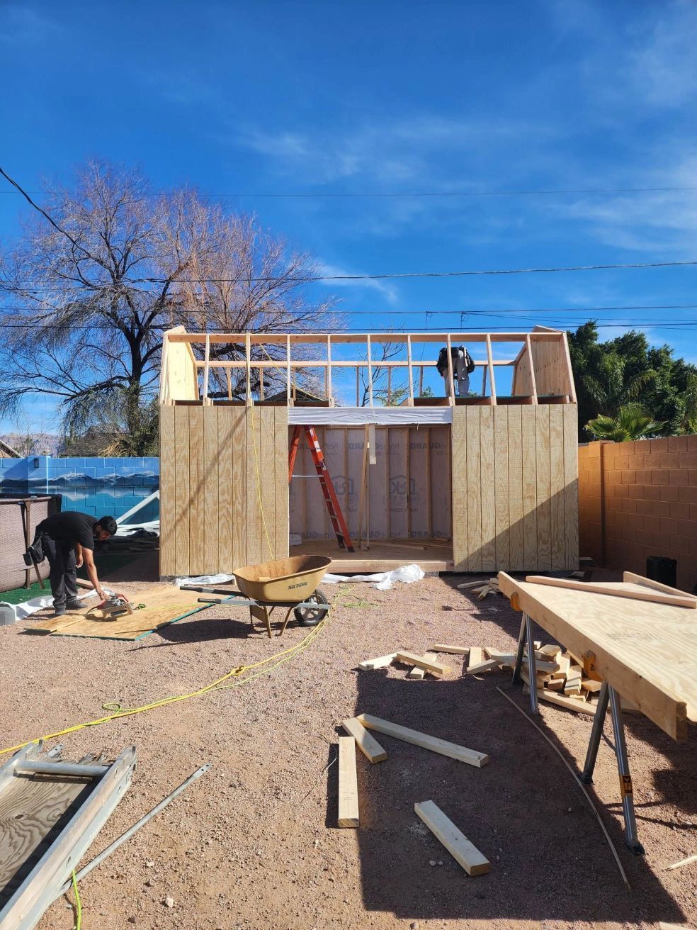 Construction site with framed wall under blue sky, tools and lumber scattered on gravel.