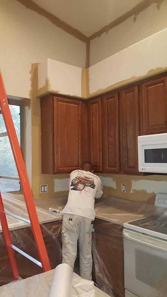 Person standing on a ladder in a kitchen, reaching up near the ceiling above brown cabinets.
