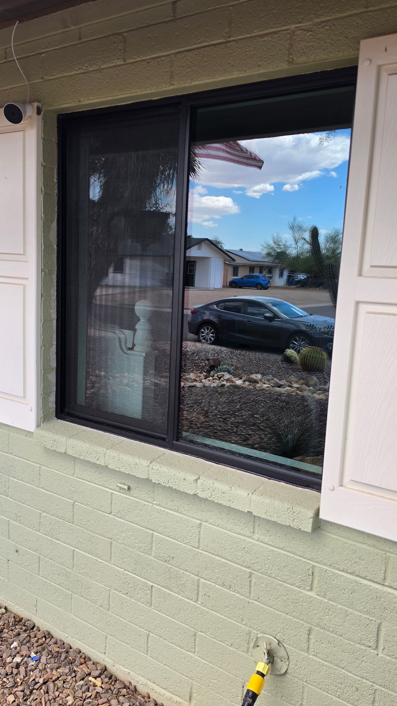 Black-framed window with white shutters reflecting a parked car and cloudy sky on a light brick wall.